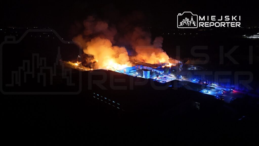 Pożar w przemysłowym obiekcie, z intensywnymi płomieniami i dymem unoszącym się w nocy, otoczony przez radiowozy.