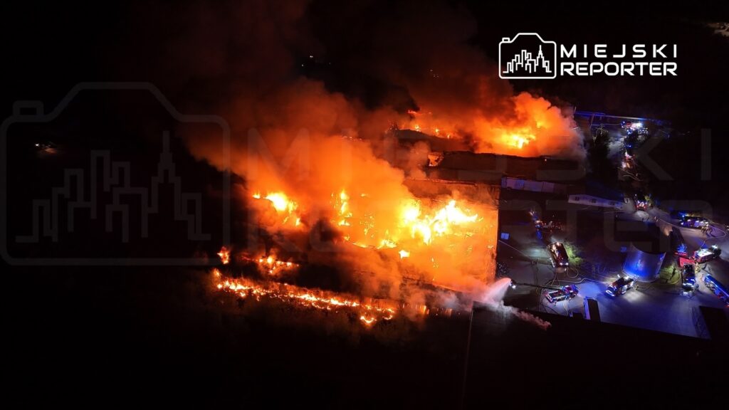 Płonący budynek otoczony przez gęsty dym, z wozami strażackimi w tle, w nocy.