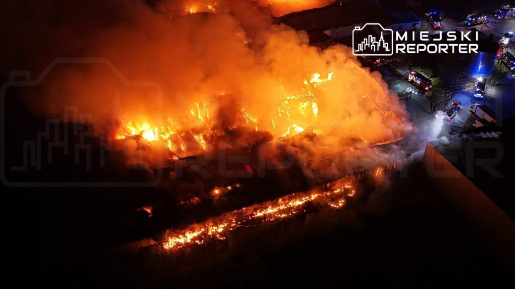 Intensywny pożar trawi budynek, z którego wydobywa się gęsty dym, w otoczeniu wozów strażackich.