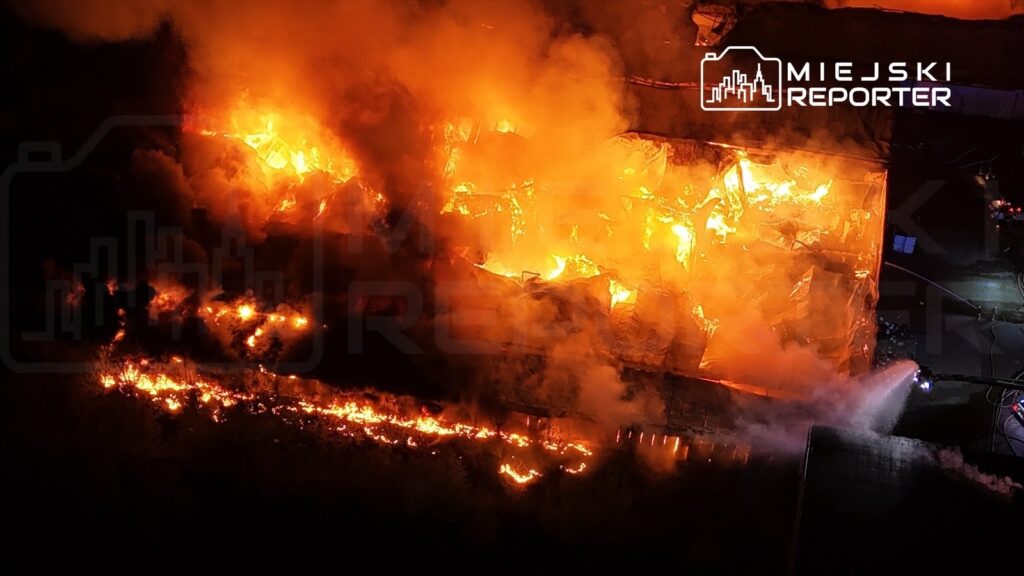Ogień engulfujący budynek, w tle widoczna akcja gaśnicza z użyciem węża strażackiego.