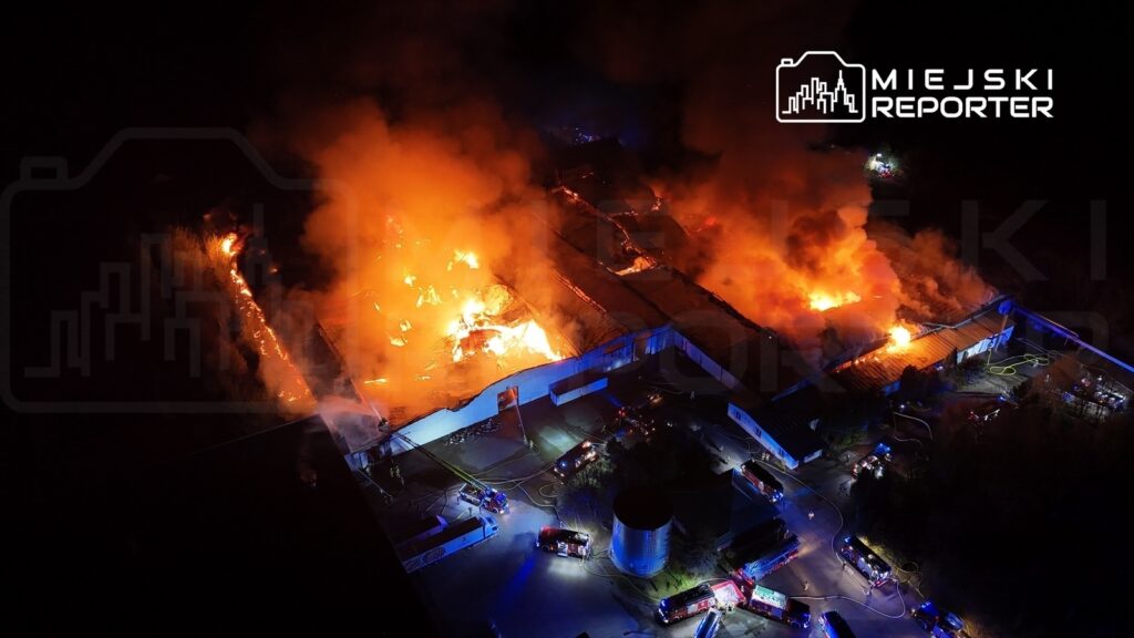 Pożar dużego budynku przemysłowego, z płomieniami i gęstym dymem w nocy, w otoczeniu wozów strażackich.