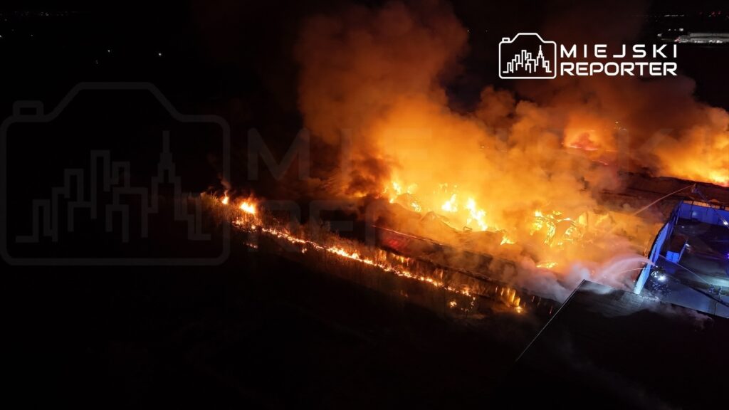 Ogromne płomienie ognia trawią zniszczony budynek, a w tle widoczny dym unoszący się w nocy.