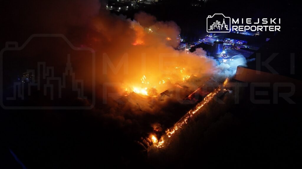 Pożar dużego obiektu przemysłowego z intensywnym dymem i płomieniami w nocy, otoczony przez pojazdy straży pożarnej.