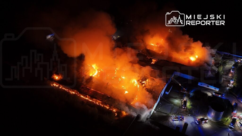 Wielki pożar strawił halę produkcyjną, generując intensywne płomienie i gęsty dym, w otoczeniu pojazdów straży pożarnej.