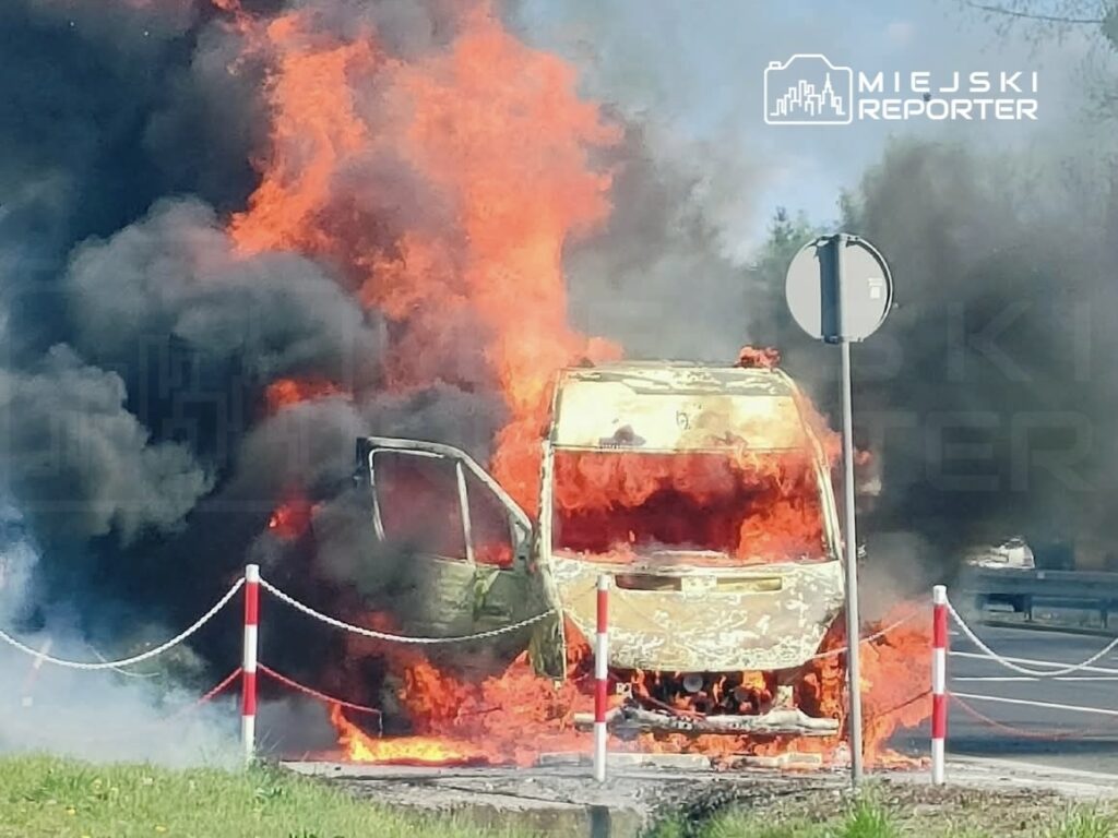 Pojazd w ogniu na poboczu drogi, otoczony gęstym dymem i płomieniami, z widocznym znakiem drogowym w tle.
