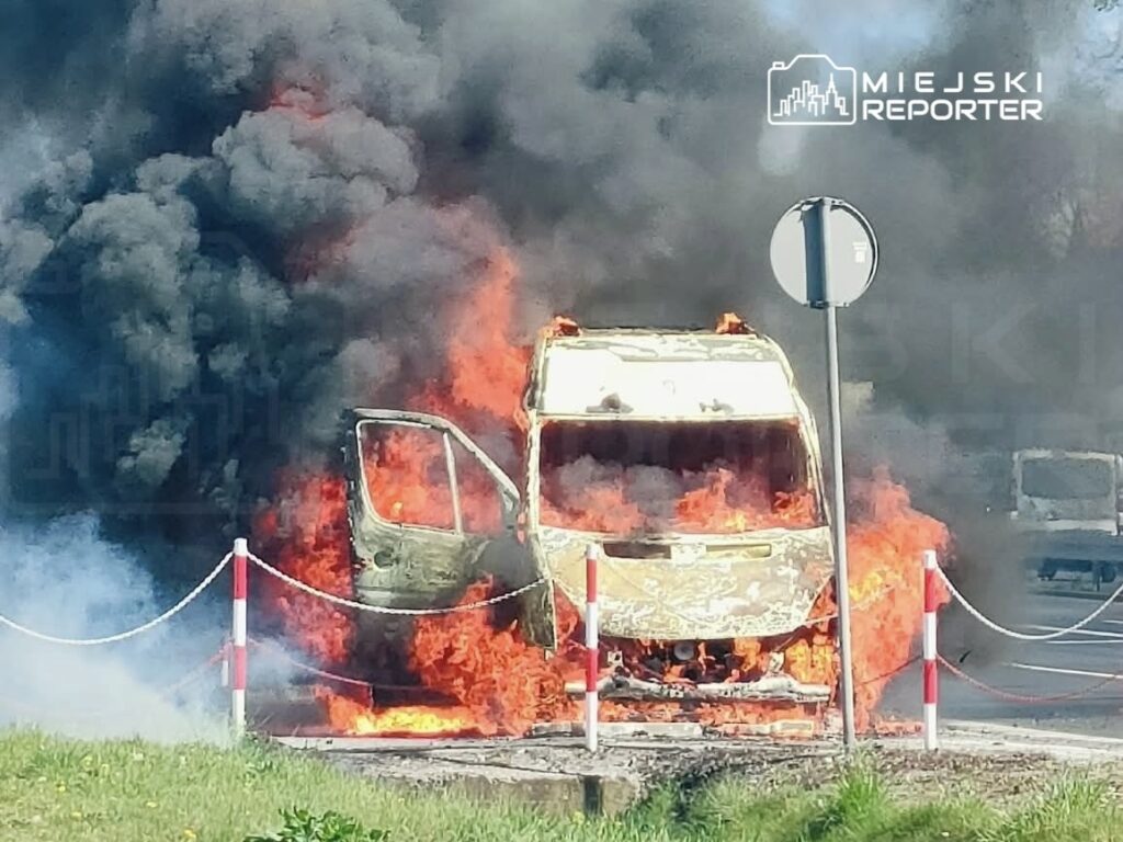 Pojazd w płomieniach na drodze, otoczony gęstym dymem, z widocznymi uszkodzeniami i przewróconym znakiem drogowym.