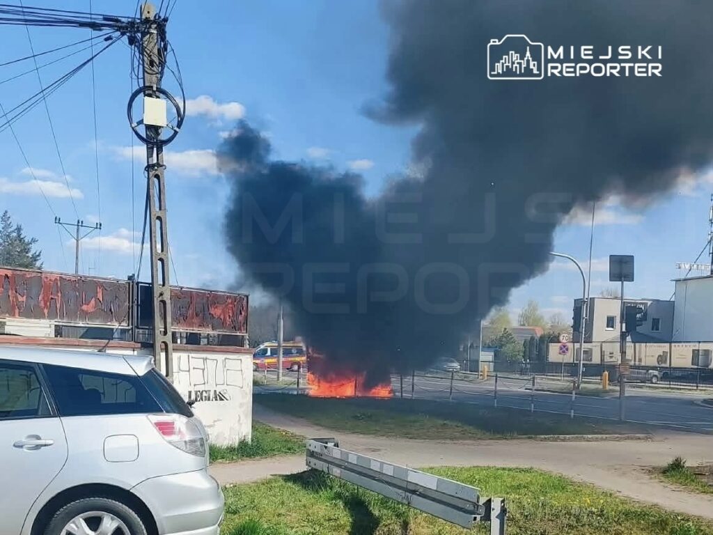 Pojazd płonący na poboczu drogi, wydobywający czarny dym, w tle widoczne budynki i znaki drogowe.