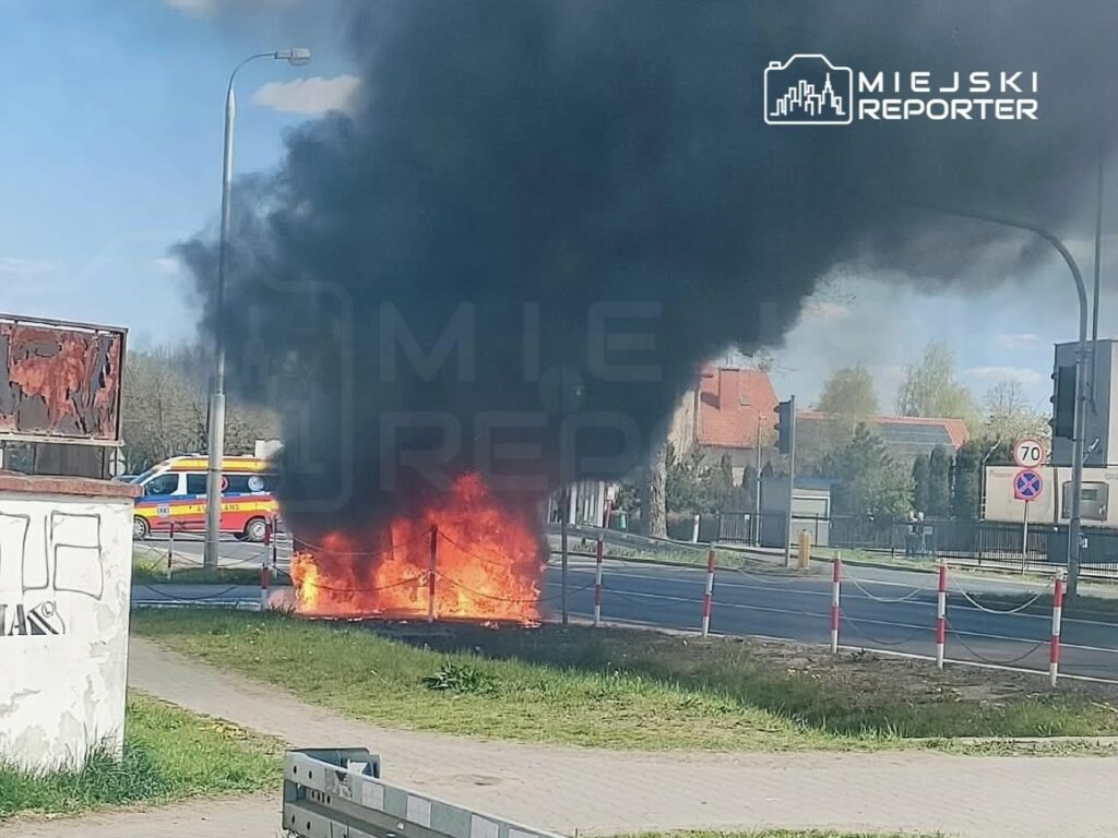 Ogień i czarny dym unoszą się z płonącego obiektu przy ruchliwej drodze, obok widoczny ambulans.