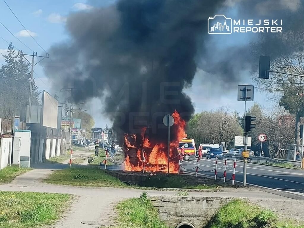 Pojazd płonący na poboczu drogi, obok strażacy gaszący ogień, w tle ruch uliczny.