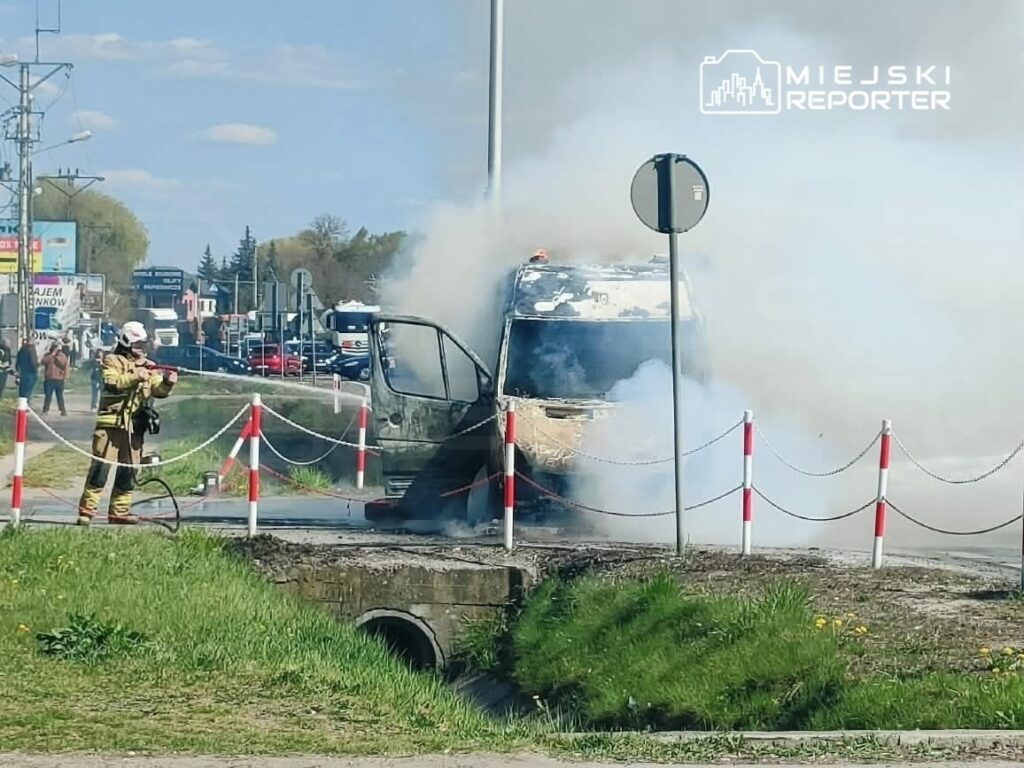 Strażak w mundurze gaśniczym gasi płonący pojazd na poboczu drogi, otoczony gęstym dymem.