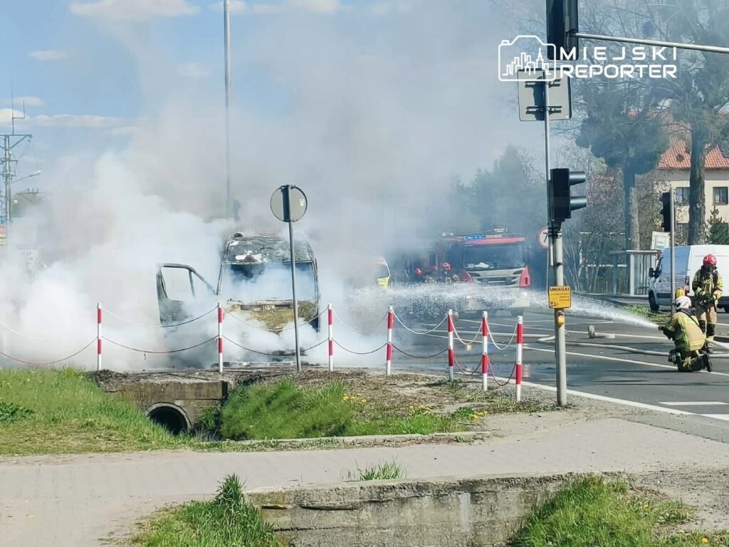 Samochód objęty dymem pożarowym, obok strażacy gaszący ogień, w tle sygnalizacja świetlna i budynki.