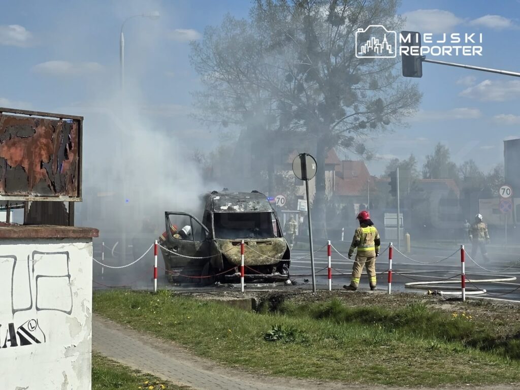 Pojazd w ogniu otoczony przez strażaków, którzy gaszą płomienie w dymiącym otoczeniu.