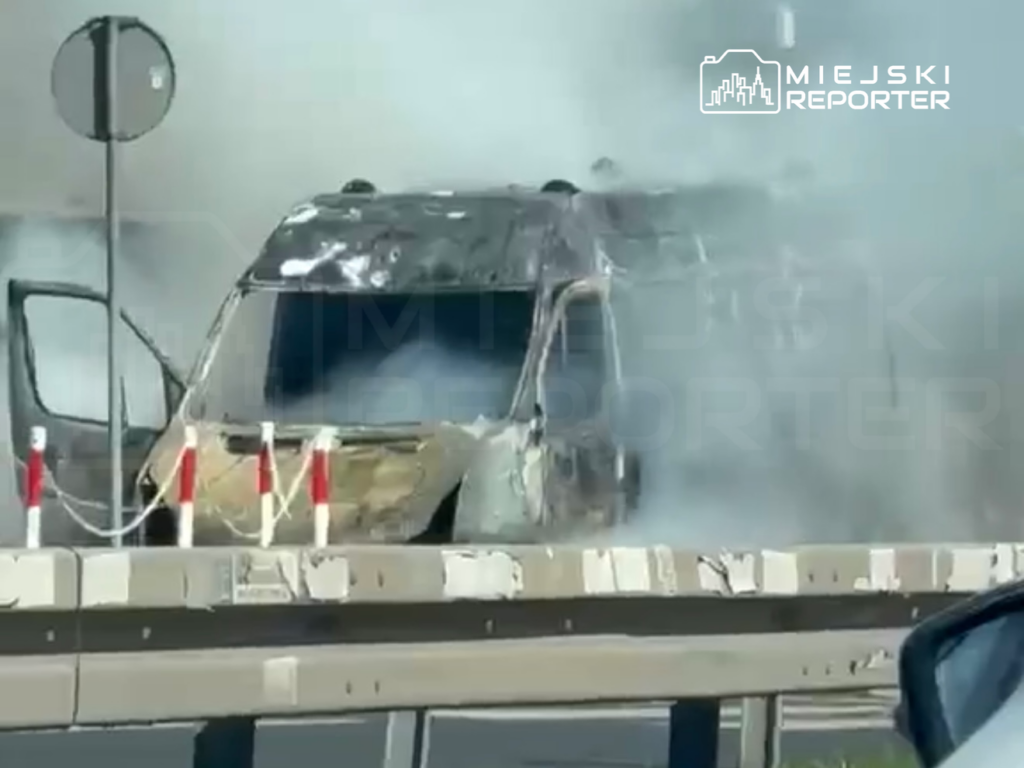 Spalony pojazd z otwartymi drzwiami, otoczony gęstym dymem, na drodze z barierkami ochronnymi.