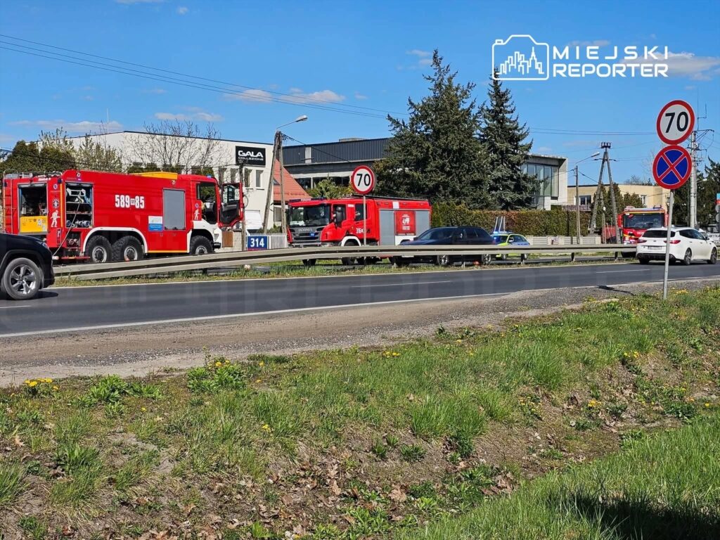 Czerwone wozy strażackie zaparkowane przy drodze, obok znaków ograniczenia prędkości i budynków.