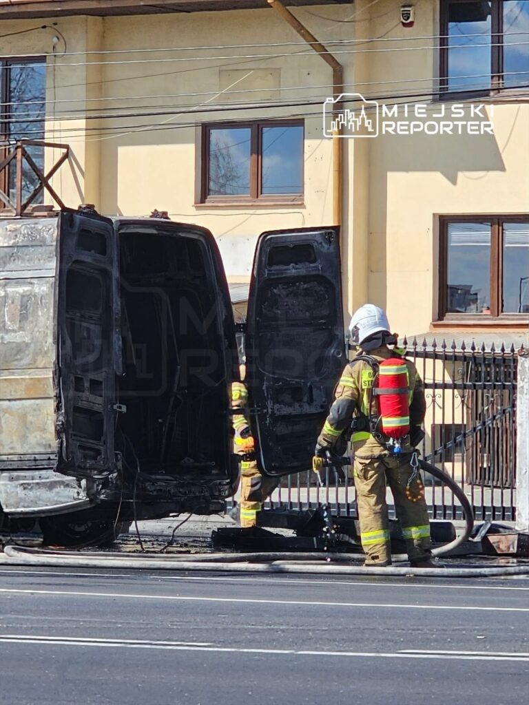 Strażacy w mundurach gaszą palący się pojazd na drodze, obok budynku mieszkalnego.
