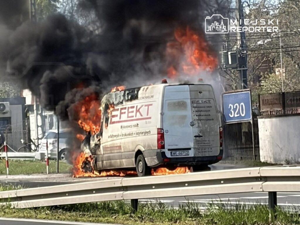 Furgonetka z płonącym przodem stoi na poboczu drogi, otoczona gęstym dymem i ogniem. W tle widoczny znak drogowy.