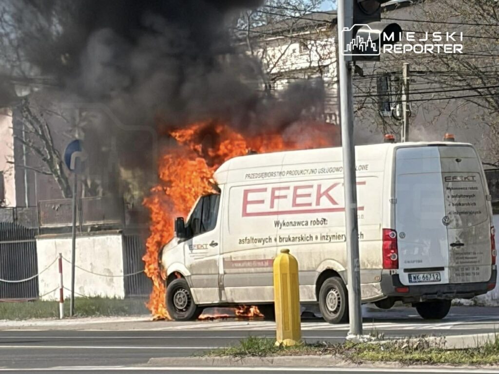 Furgonetka z logo "EFKT" płonie na poboczu drogi, wydobywając intensywny dym i ogień.