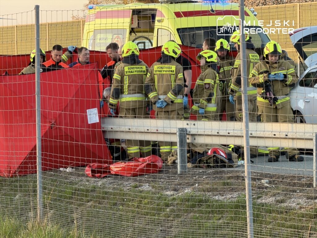 Grupa strażaków w odblaskowych ubraniach pracuje przy wypadku drogowym, otoczona czerwonymi zasłonami.