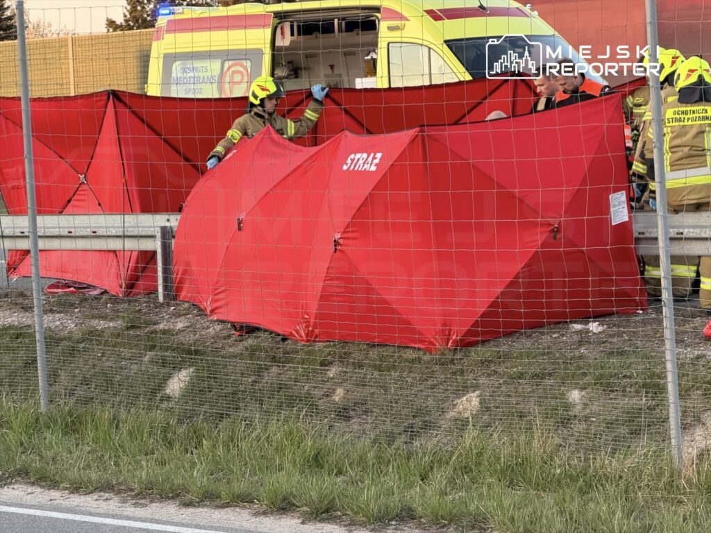 Strażacy pracują przy czerwonych namiotach rozstawionych obok drogi, w tle widoczna karetka pogotowia.