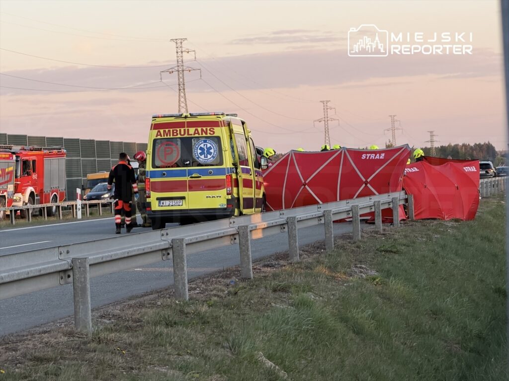 Zespół ratunkowy ustawia namioty ochronne obok karetki na drodze, w tle widoczne pojazdy straży pożarnej.