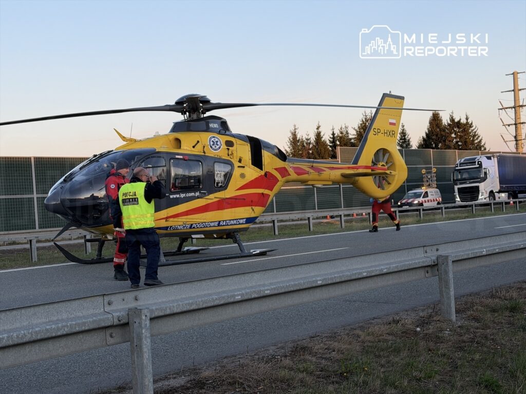 Ratownicy medyczni rozmawiają obok helikoptera Lotniczego Pogotowia Ratunkowego na drodze, w tle widoczne ciężarówki.