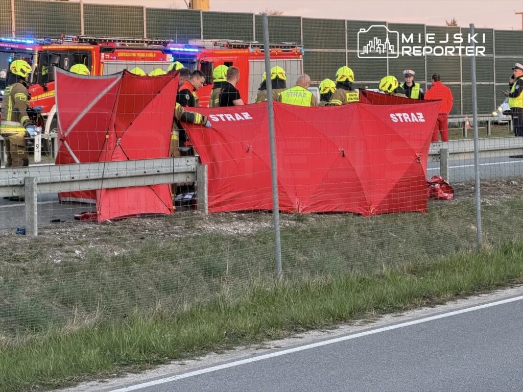 Grupa strażaków pracuje przy wypadku drogowym, otoczona czerwonymi namiotami, na poboczu drogi.
