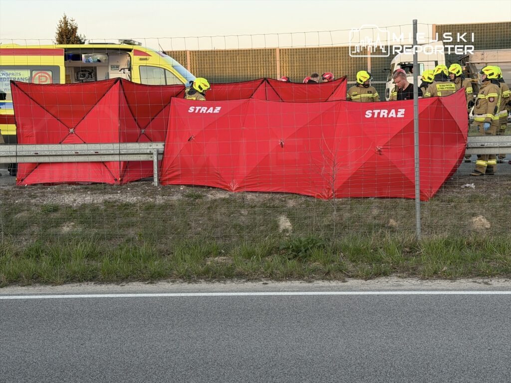 Grupa strażaków w żółtych hełmach pracuje przy czerwonych namiotach ratunkowych obok drogi, w tle widać ambulans.