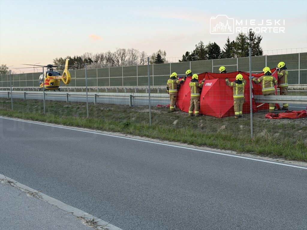 Grupa strażaków ustawia czerwoną osłonę wzdłuż drogi, obok lądowania helikoptera ratunkowego.