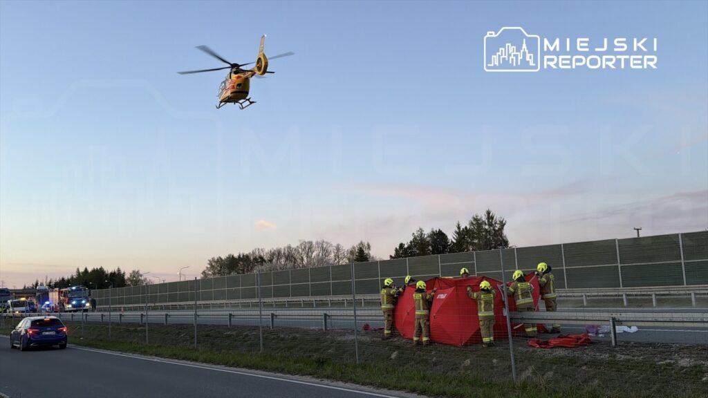 Helikopter ratunkowy przelatuje nad autostradą, gdzie strażacy przygotowują namiot medyczny obok oznakowania akcji ratunkowej.