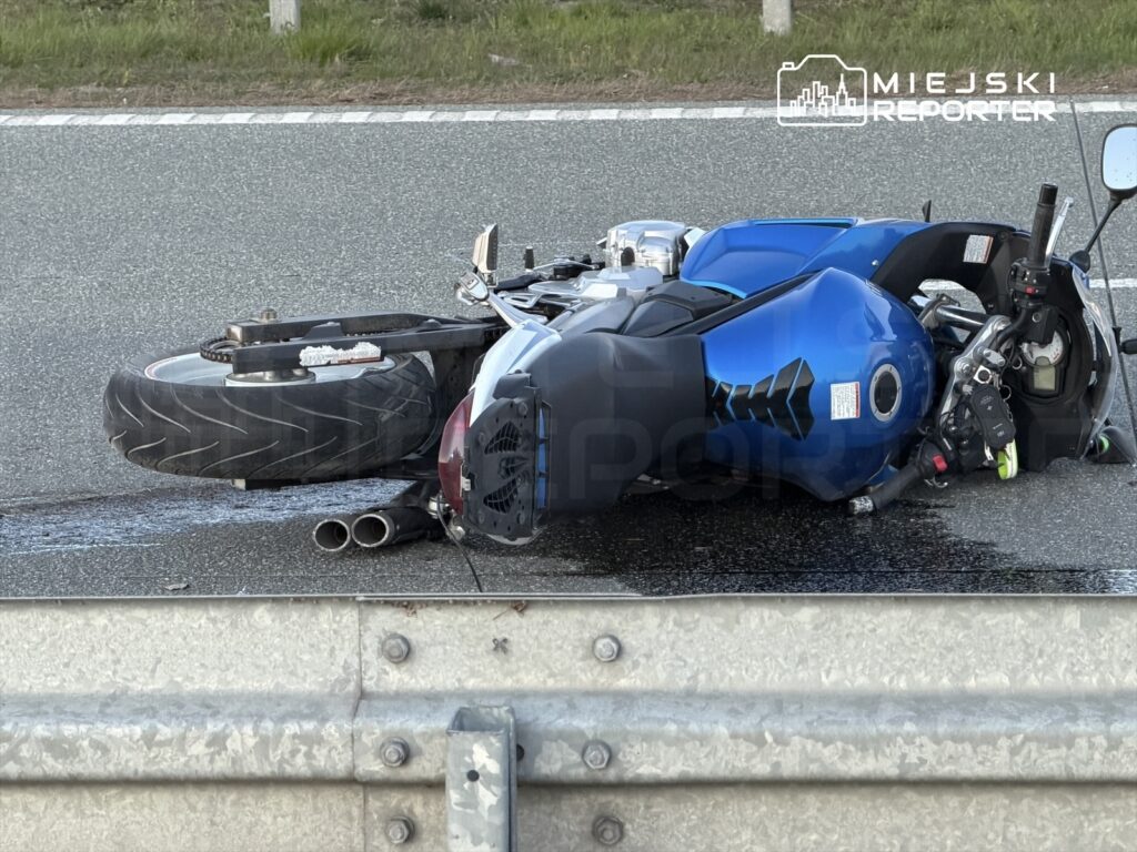 Motocykl leżący na drodze, z uszkodzoną przednią częścią, obok barierki ochronnej.