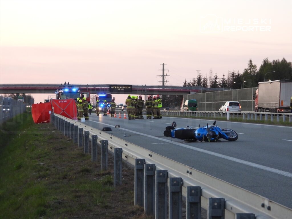 Grupa strażaków pracuje przy wypadku drogowym na autostradzie, obok leżącego niebieskiego motocykla.
