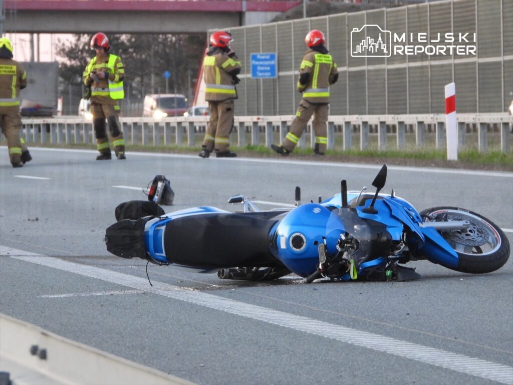Motocykl leżący na drodze, obok strażacy w mundurach, monitorujący sytuację na autostradzie.