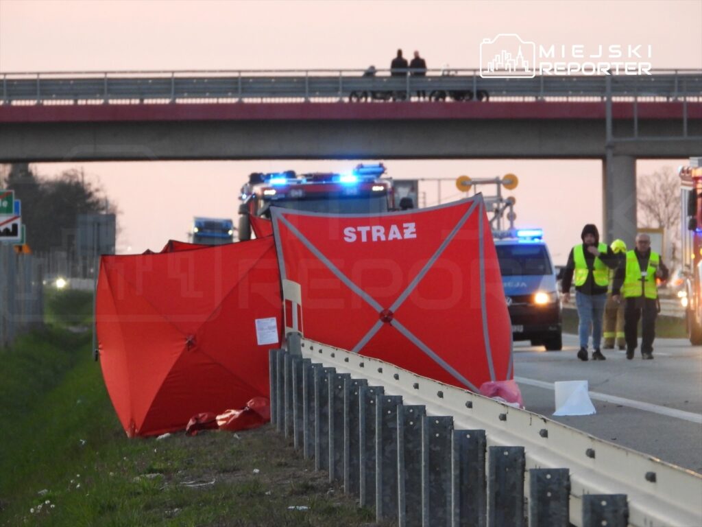 Na poboczu drogi rozstawione są czerwone namioty z napisem "STRAŻ", a w tle widać pojazdy ratunkowe.
