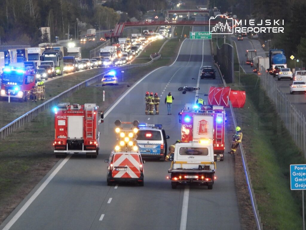 Na drodze stoi kilka pojazdów ratunkowych, a strażacy pracują przy wypadku, obok leży motocykl.