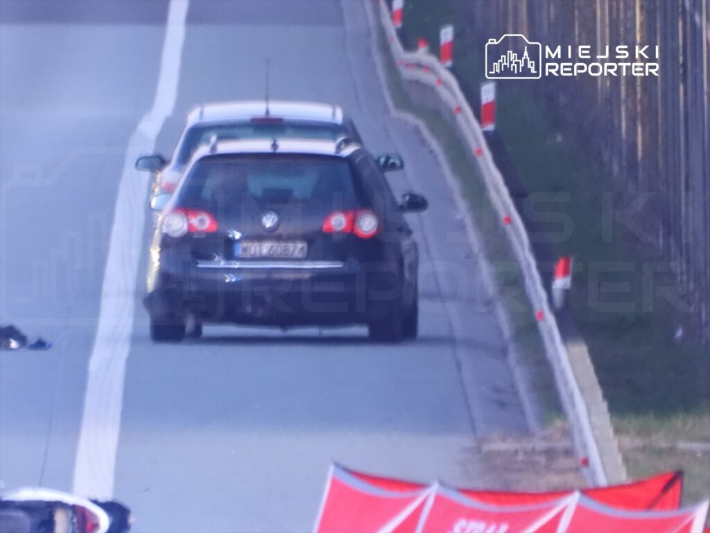 Czarny samochód osobowy porusza się po drodze, obok leżają fragmenty odzieży i znaki ostrzegawcze.