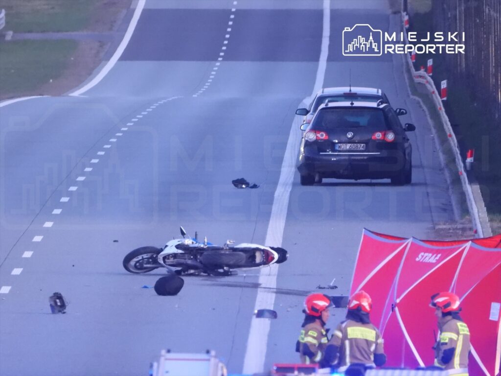 Motocykl leży na jezdni, obok rozrzucone części. W tle widać samochód oraz grupę strażaków przy czerwonym namiocie.