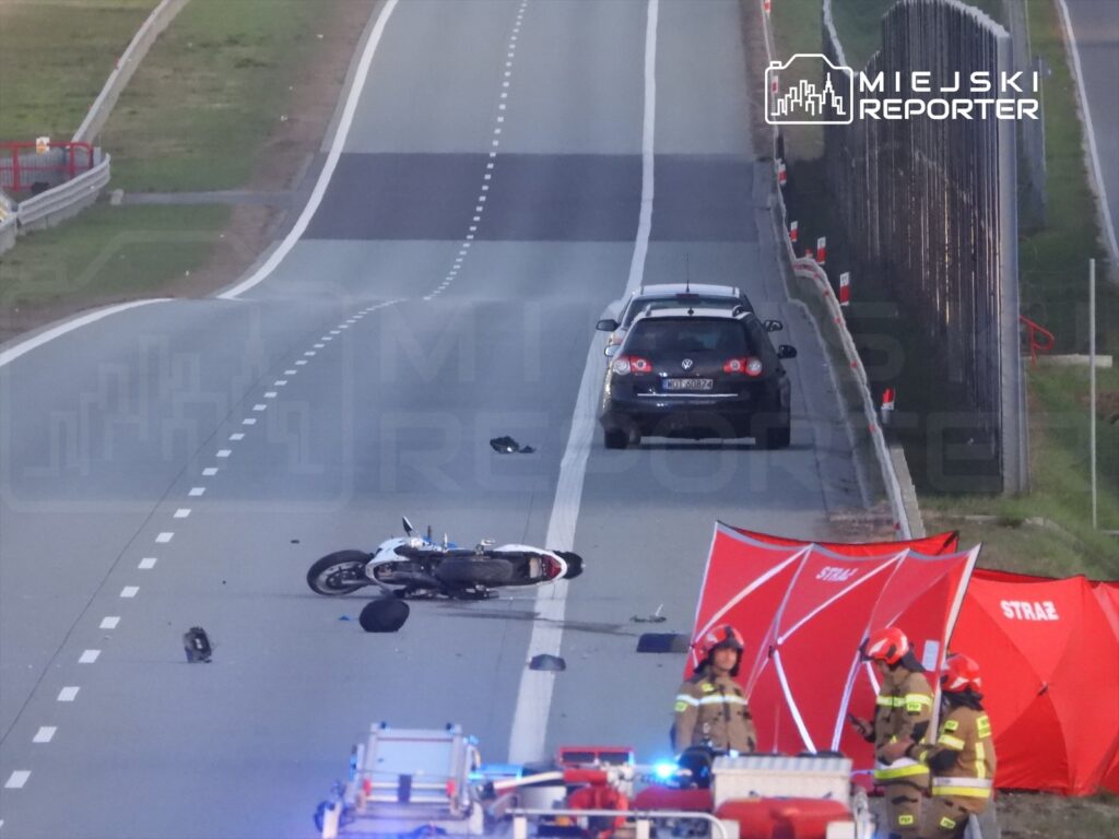 Motocykl leży na drodze obok rozbitego samochodu, a strażacy prowadzą działania ratunkowe w pobliżu.