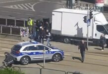 Grupa osób z interwencją policji przy białej ciężarówce na ulicy, obok przejeżdżający rowerzysta.