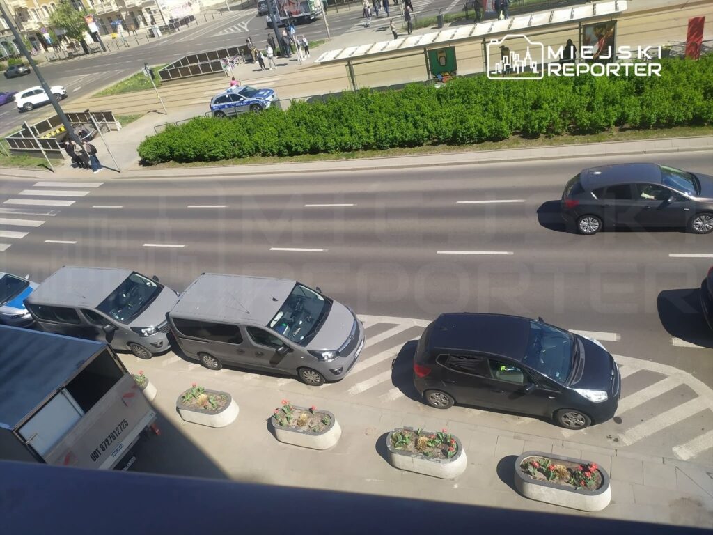 Ruch samochodowy na ulicy z zaparkowanymi pojazdami, w tle przejście dla pieszych i zieleń.