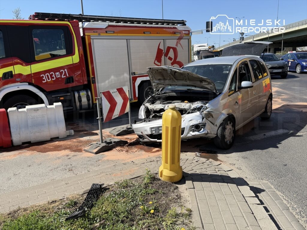 Zniszczony samochód osobowy z otwartą maską stoi obok wozu strażackiego na ruchliwej ulicy, w otoczeniu barier ochronnych.