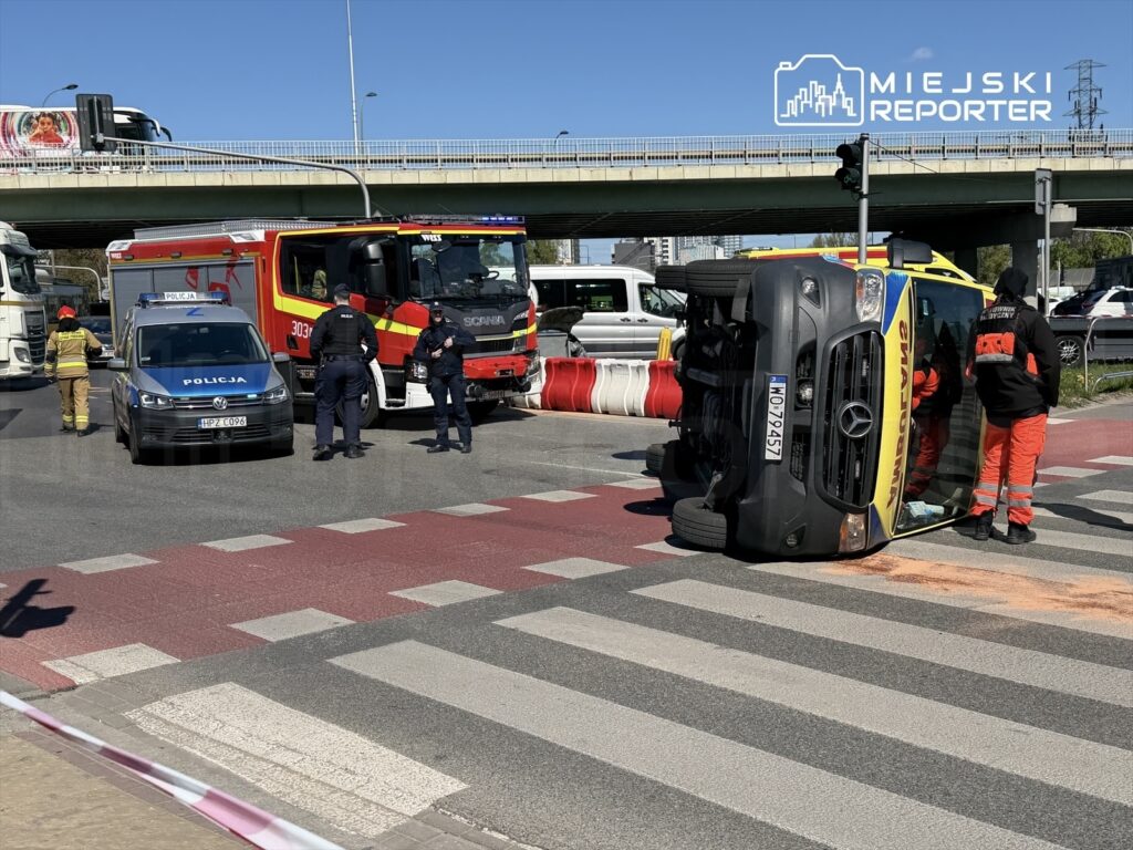 Na skrzyżowaniu leży przewrócony pojazd, obok niego interweniują strażacy i policja, a w tle widoczne inne samochody.