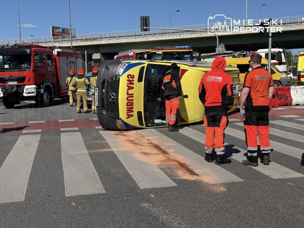 Na drodze przewrócona karetka, obok pracują strażacy i ratownicy medyczni w pomarańczowych strojach, w tle wozu strażackiego.