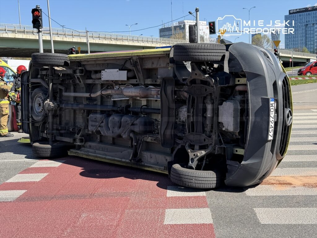Samochód osobowy marki Mercedes leży na boku na skrzyżowaniu, obok pracują strażacy w kaskach.