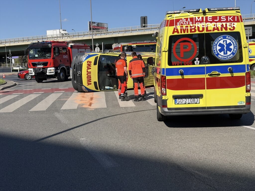 Na skrzyżowaniu leży przewrócona karetka, obok niej stoją ratownicy w czerwonych uniformach, a w tle widać wóz strażacki.