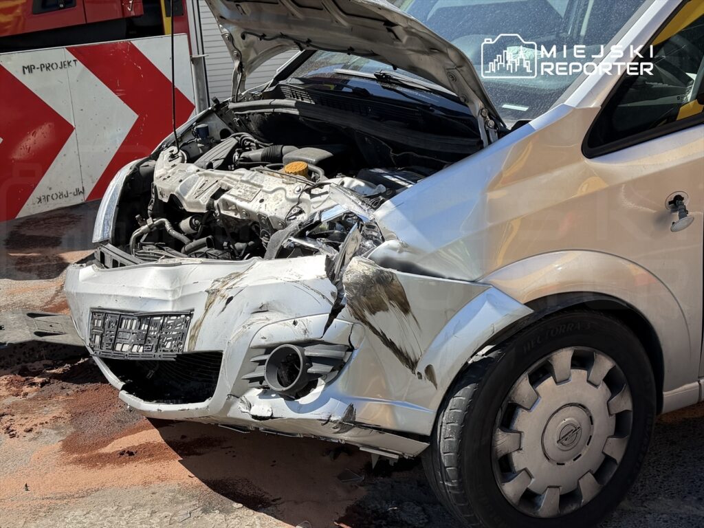 Uszkodzony srebrny samochód osobowy z otwartą maską, stojący na drodze obok pojazdu służb ratunkowych.
