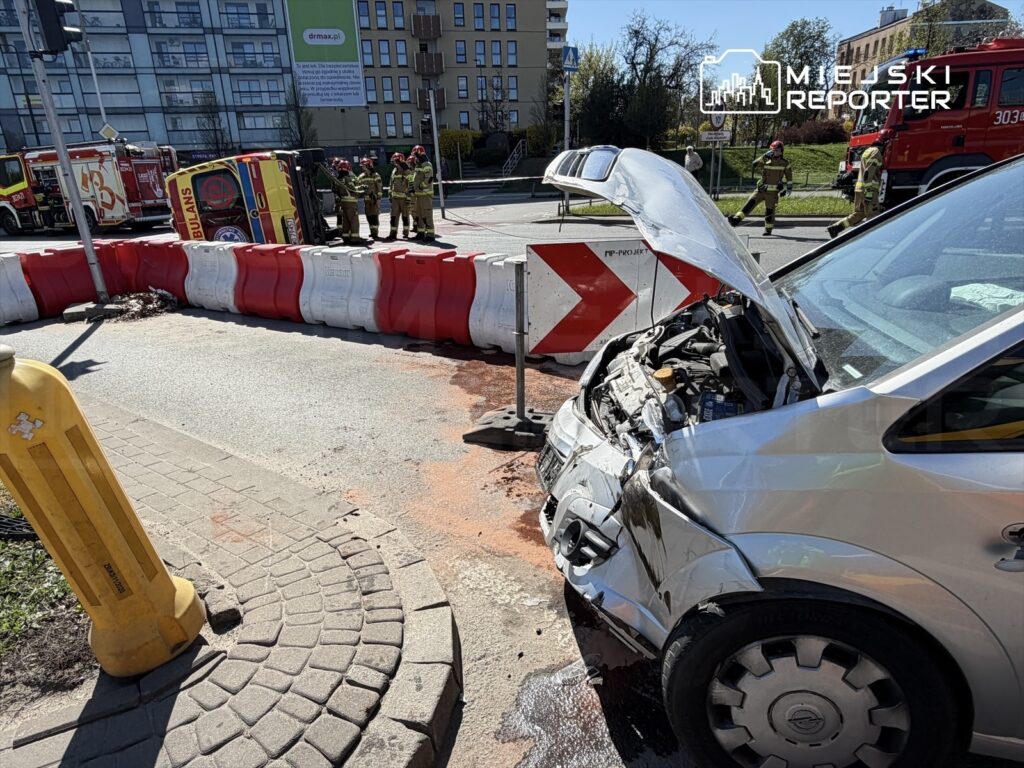 Uszkodzony samochód osobowy z otwartą maską stoi przy barierze drogowej, a w tle widać strażaków oraz wozów strażackich.
