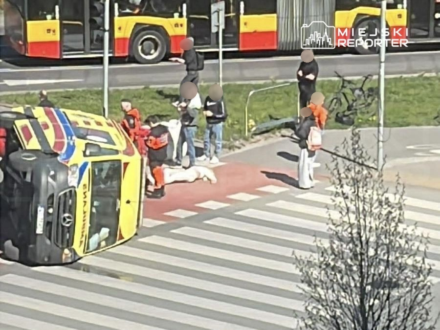 Na przewróconym pojeździe ratunkowym stoją ratownicy, a obok leży poszkodowany mężczyzna na przejściu dla pieszych.