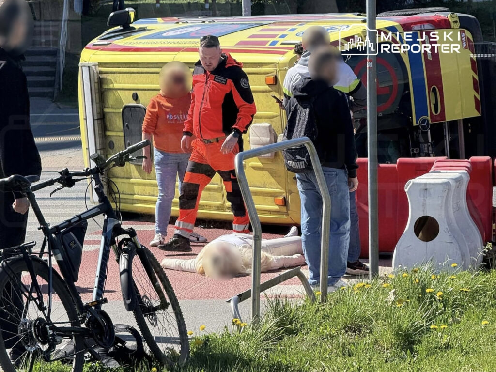 Ratownik medyczny udziela pomocy poszkodowanej osobie leżącej na jezdni obok przewróconego pojazdu.