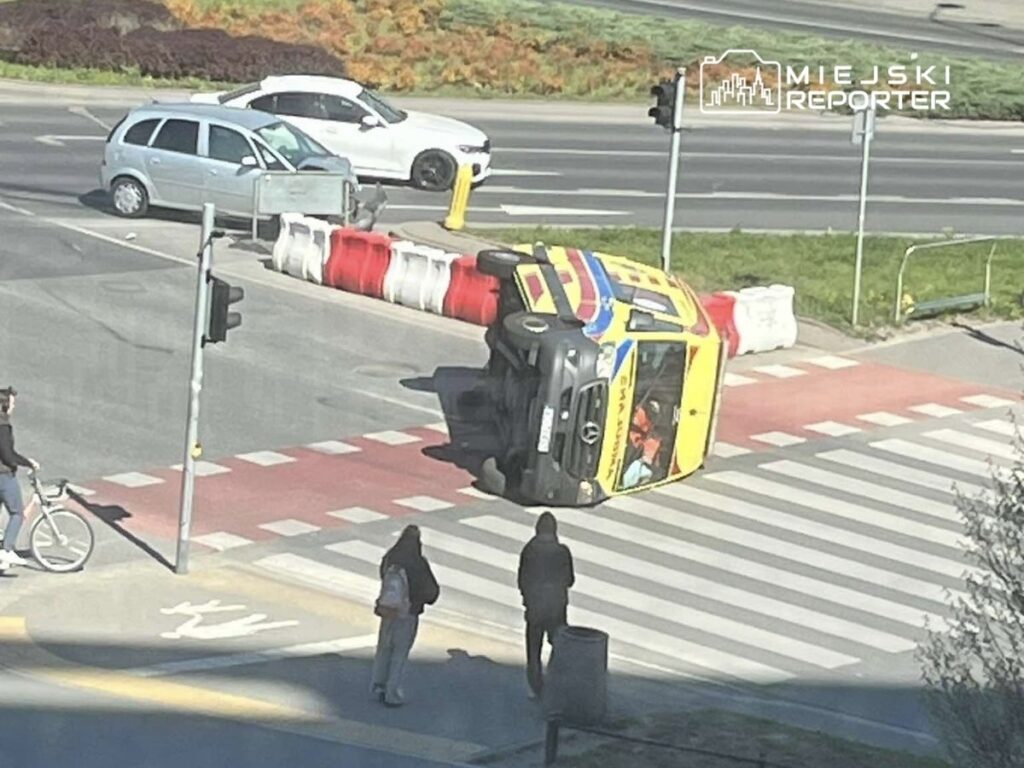 W przewróconym autobusie na skrzyżowaniu stoją dwie osoby, a w tle widać inne pojazdy oraz sygnalizację świetlną.