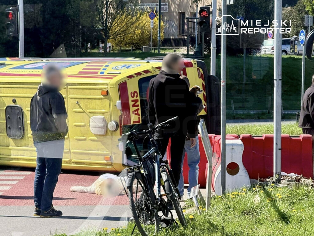 Żółta karetka leży na boku, obok grupy ludzi, z których jeden przygląda się leżącej osobie na ziemi.